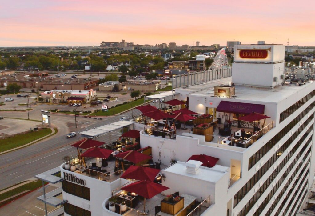 An aerial view of the Hilton Hotel with the rooftop bar, Reveille, featuring red umbrellas and seating areas. In the background, the city stretches out in the distance with roads, cars, and surrounding buildings visible. Above, the sky glows with vivid pink and orange streaks from the sunset, creating a striking backdrop over the urban landscape.